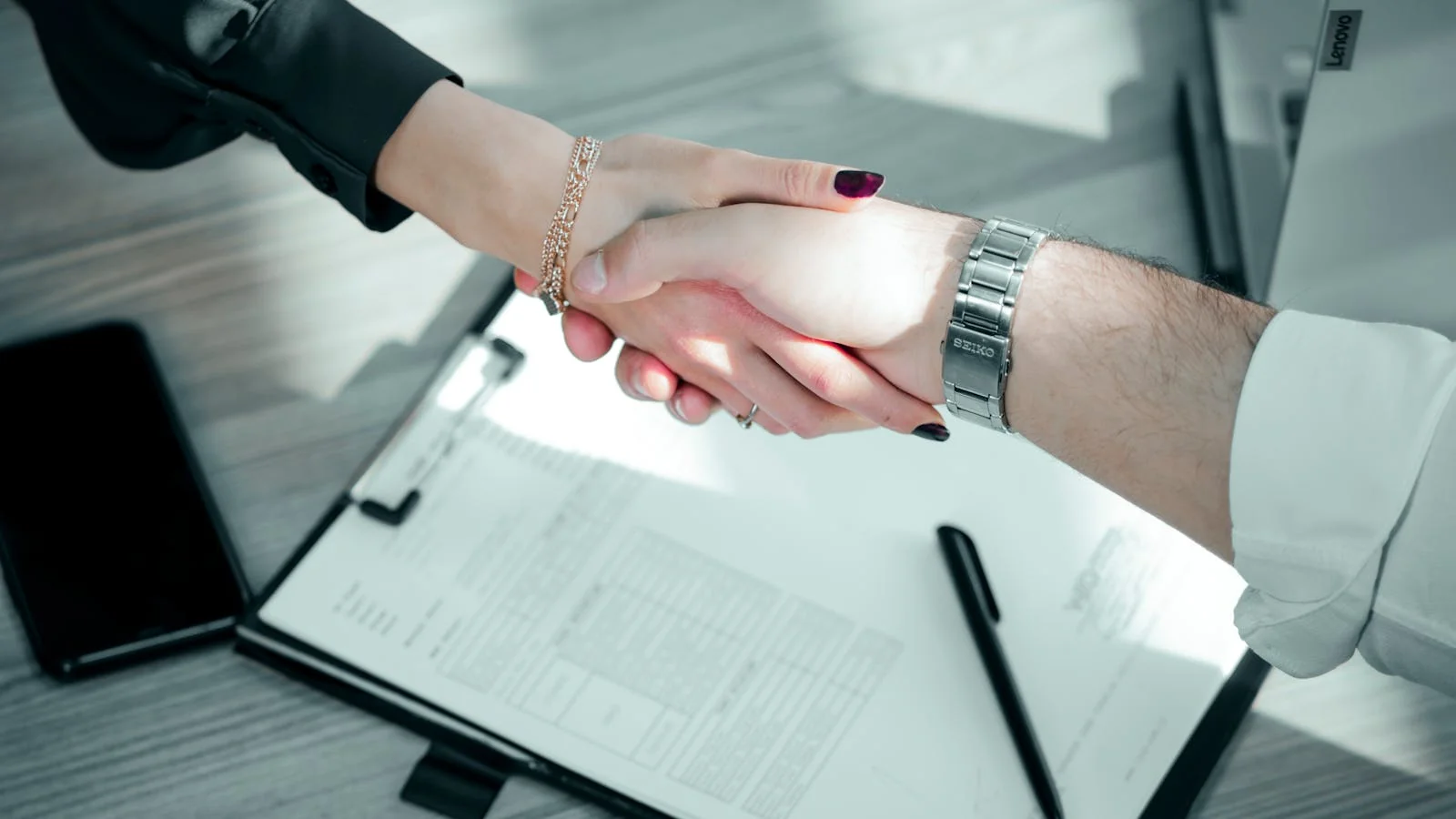 A close-up of two professionals shaking hands across a contract on a wooden table, sealing a new PR agency retainer agreement.
